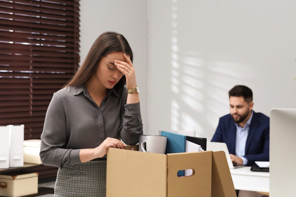 A woman with all her office items in a box, while she looks down with her hand on her forehead after losing her job.
