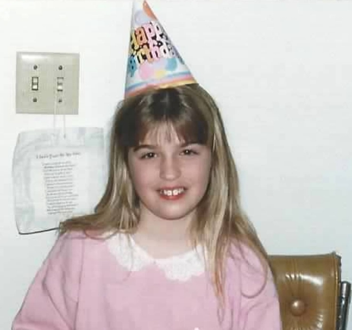 Another picture from the author's childhood where is in a pink top with a birthday hat on.