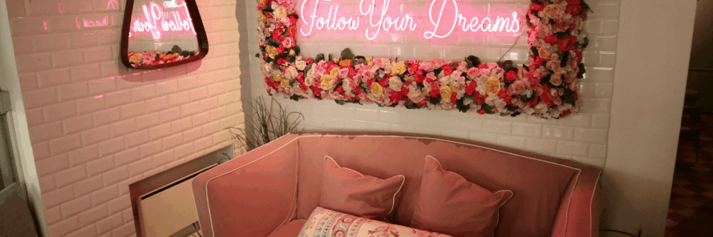 A mirror surrounded by flowers with a neon light that says follow your dreams hangs on an ivory colored wall. There is a pink couch.