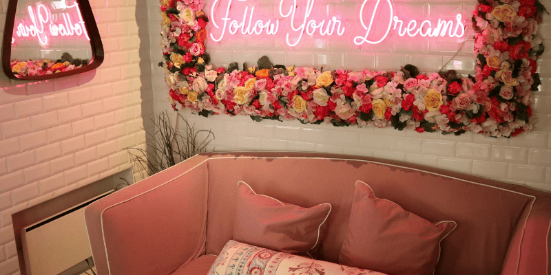 A mirror surrounded by flowers with a neon light that says follow your dreams hangs on an ivory colored wall. There is a pink couch.