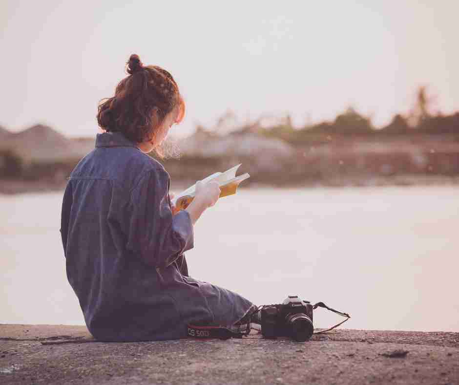 Photo of a girl sitting by the water reading a book with a camera next to her