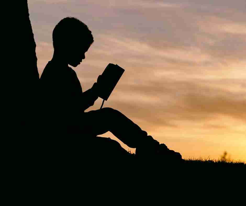 Photo of a child silhouetted against sunset, leaning on tree reading a book