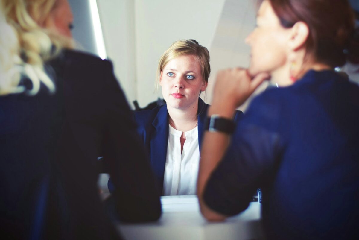 Three career women sitting at a table in a meeting. The woman facing us appears deep in thought signifying a career challenge.