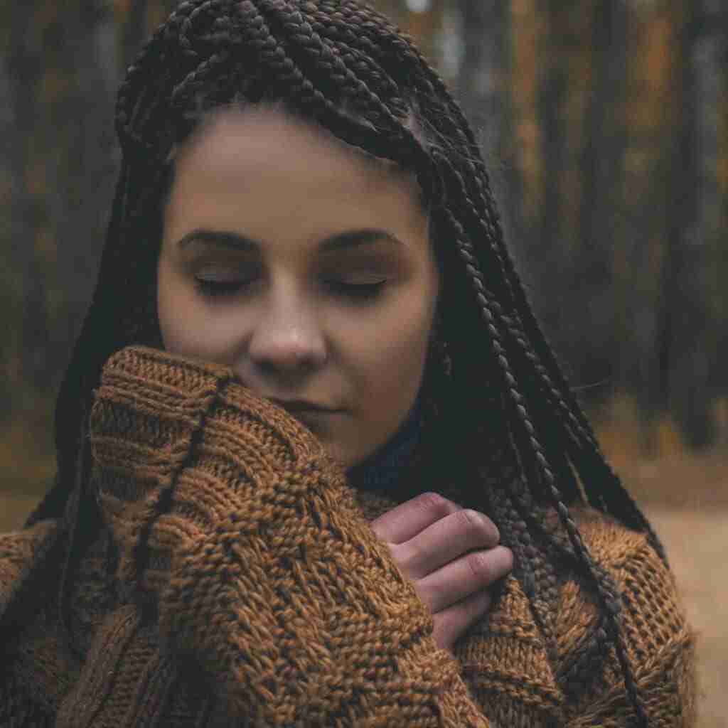 Black/Indigenous woman with long dark braids and cozy sweater in woods eyes closed looking peaceful and happy practicing mindfulness