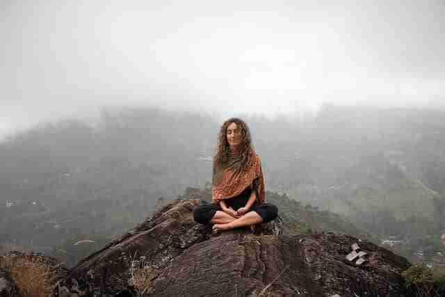 Woman sitting on a rocky hill in front of a cloud-shrouded mountain practicing mindfulness