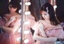 Asian woman dressed in pink dress looking in mirror with bright lights all around.