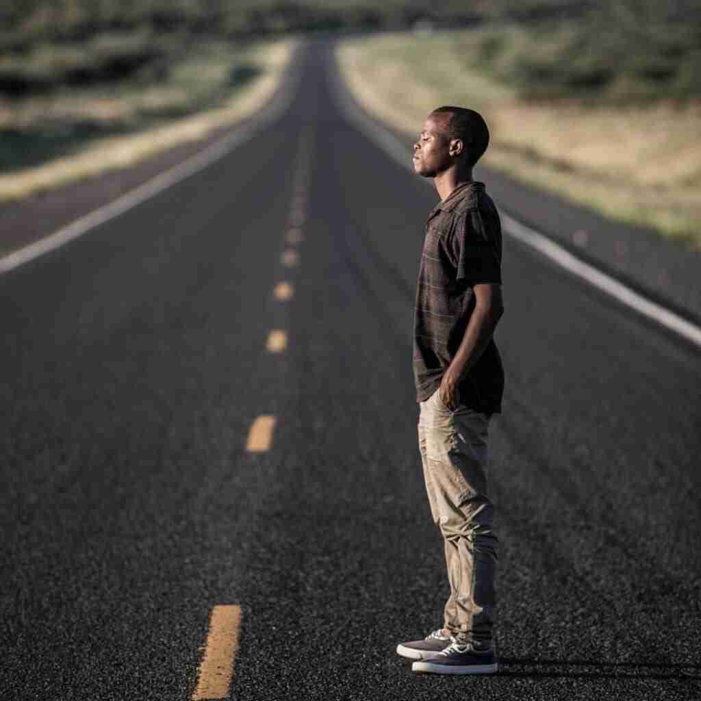 Untitled design (26) - Resilient Stories Black man standing on empty road with head up and eyes closed, facing west toward sun.