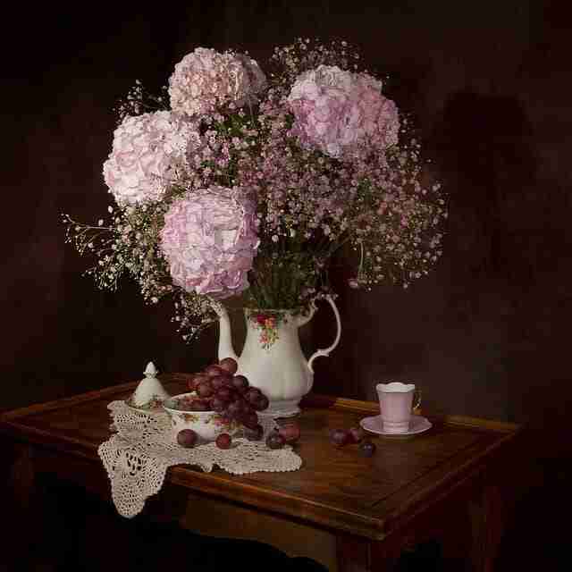 antique teapot with flower arrangement of pink hydrangeas and pink baby's breath on a table with tea cups and grapes