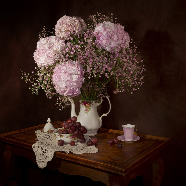 antique teapot with flower arrangement of pink hydrangeas and pink baby's breath on a table with tea cups and grapes 