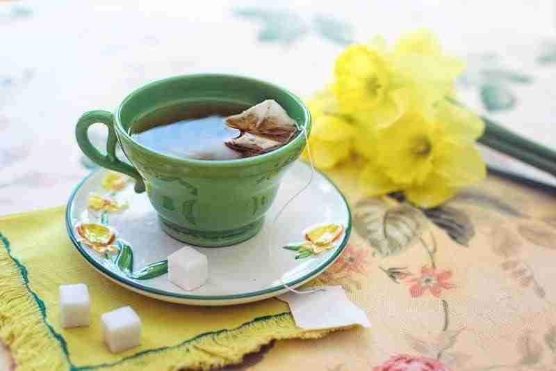Untitled-design-2024-05-20T153557.449 - Resilient Stories Green teacup on a ceramic saucer with steeping herbal tea inside, a few sugar cubes and a bouquet of daffodils sit next to the cup.