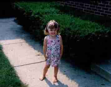 Kristin Oesterle as a very young girl, standing in front of shrubbery of a home in a floral jumper.