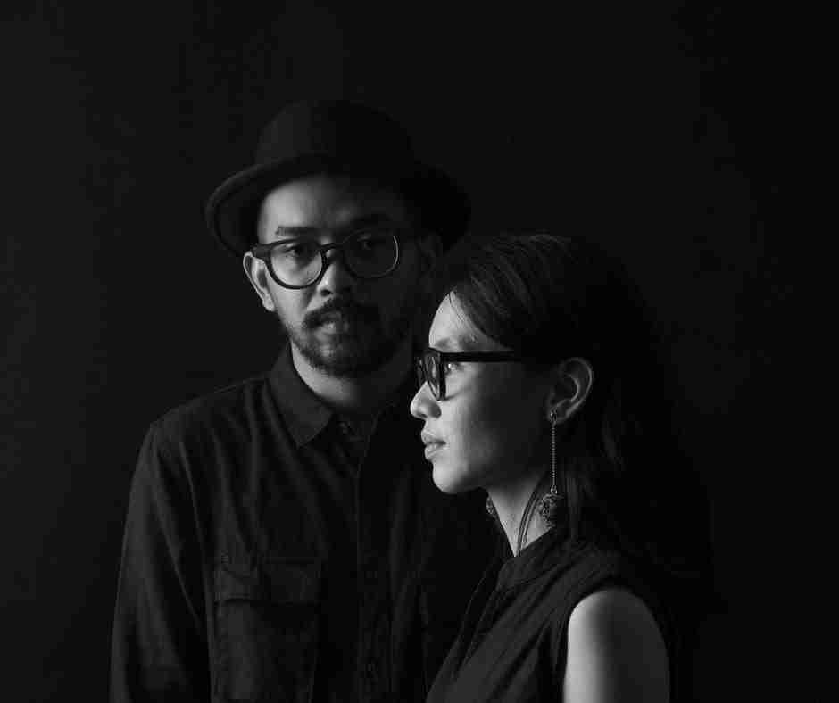 Black and white image of man and woman, both wearing glasses, standing perpendicular to one another, looking somber