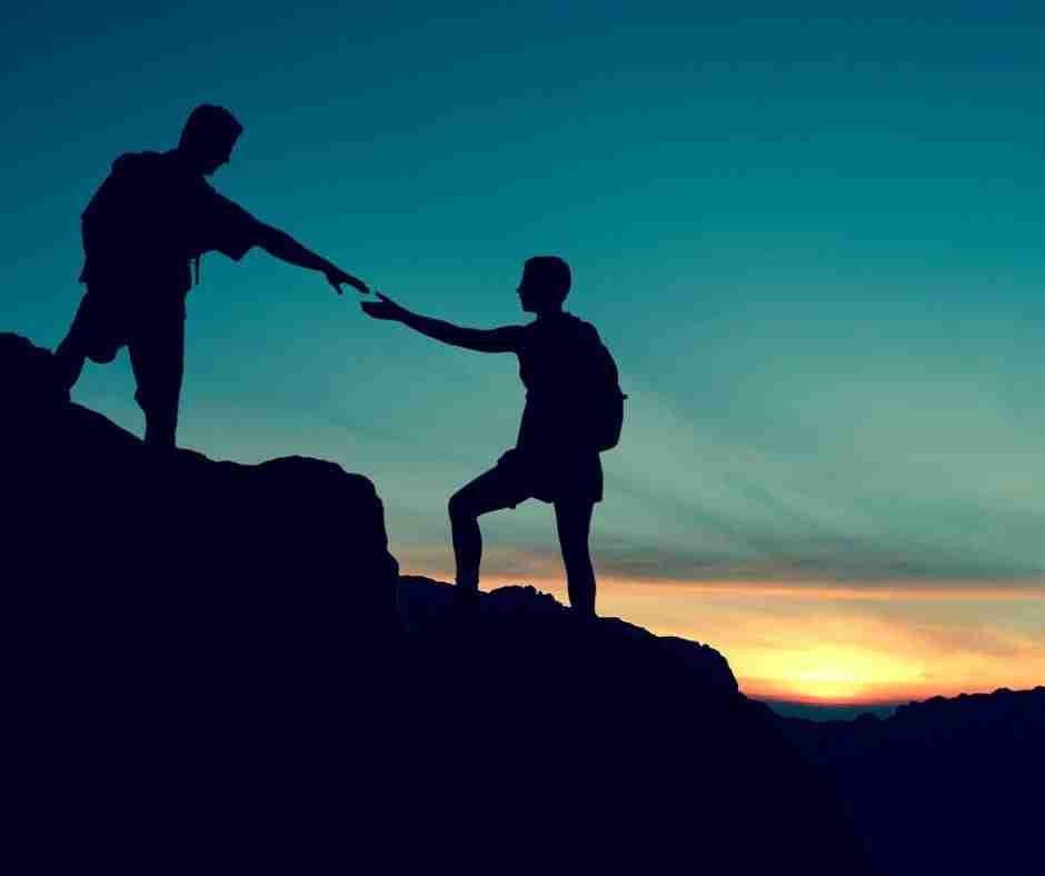 Untitled design (22) - Resilient Stories Two silhouetted mountain climbers, one outstretching a hand to thelp the other, in front of a beautiful sunset.