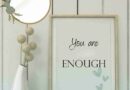 A pretty wall art in soft green that says you are enough with a mirror on the wall and a vase of soft greenery.