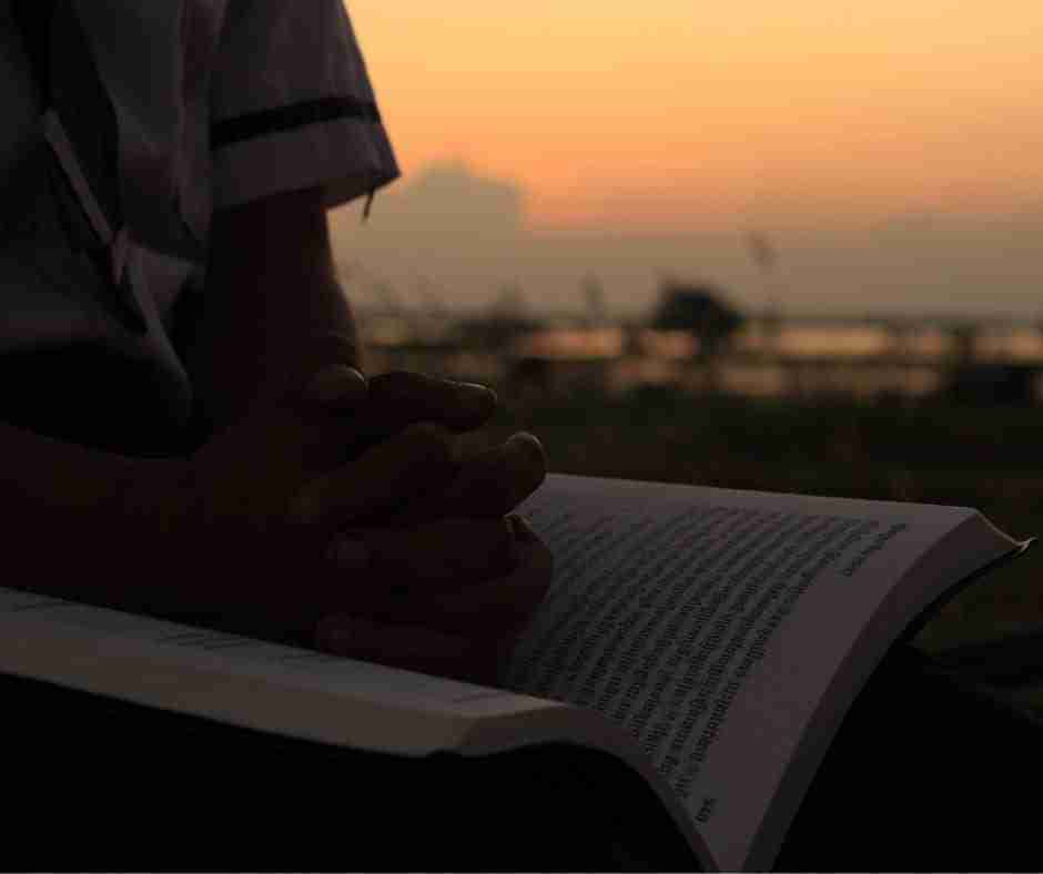 Person with hands folded over a book with Sanskrit writing, sitting in front of a dusty sunset.