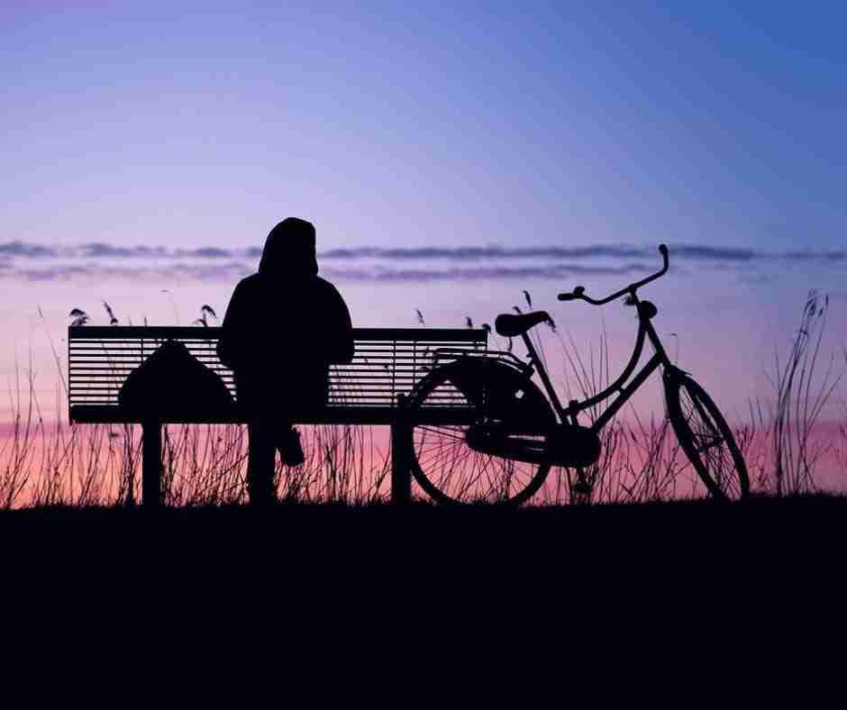 Photo of silhouette of person on park bench with a bicylce parked next to them, watching a beautiful purple and pink sunset in the sky.