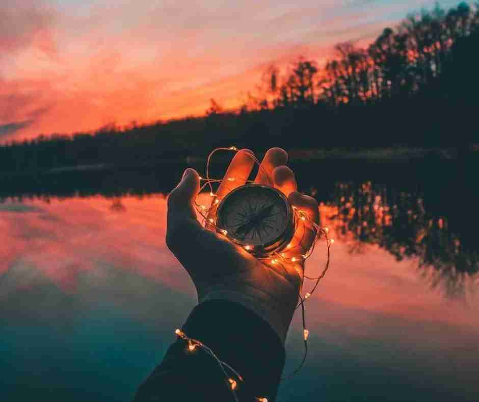 Lighted hand holding a compass over a lake with a beautiful pink sunset in the sky behind, symbolizing be true to yourself and follow your own path.
