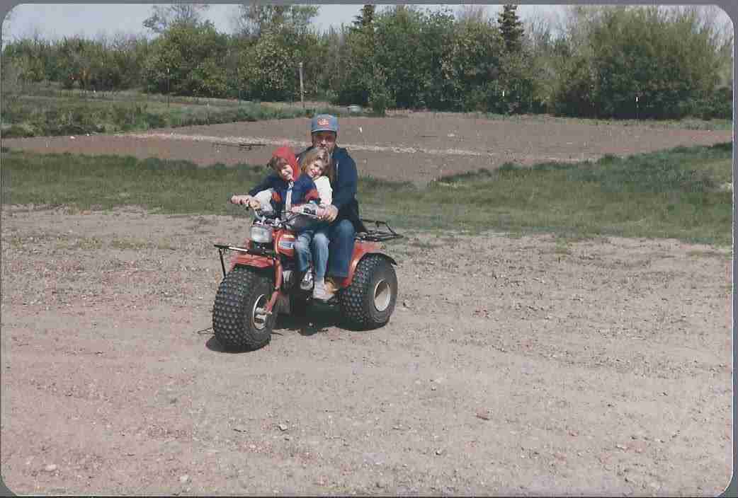 Donna and Michael as children on a three-wheeler with Grandpa at the farm.