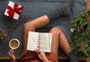 Black female in cozy socks and sweater sitting on blanket with cup of coffee and cozy book amidst holiday gifts and decorations