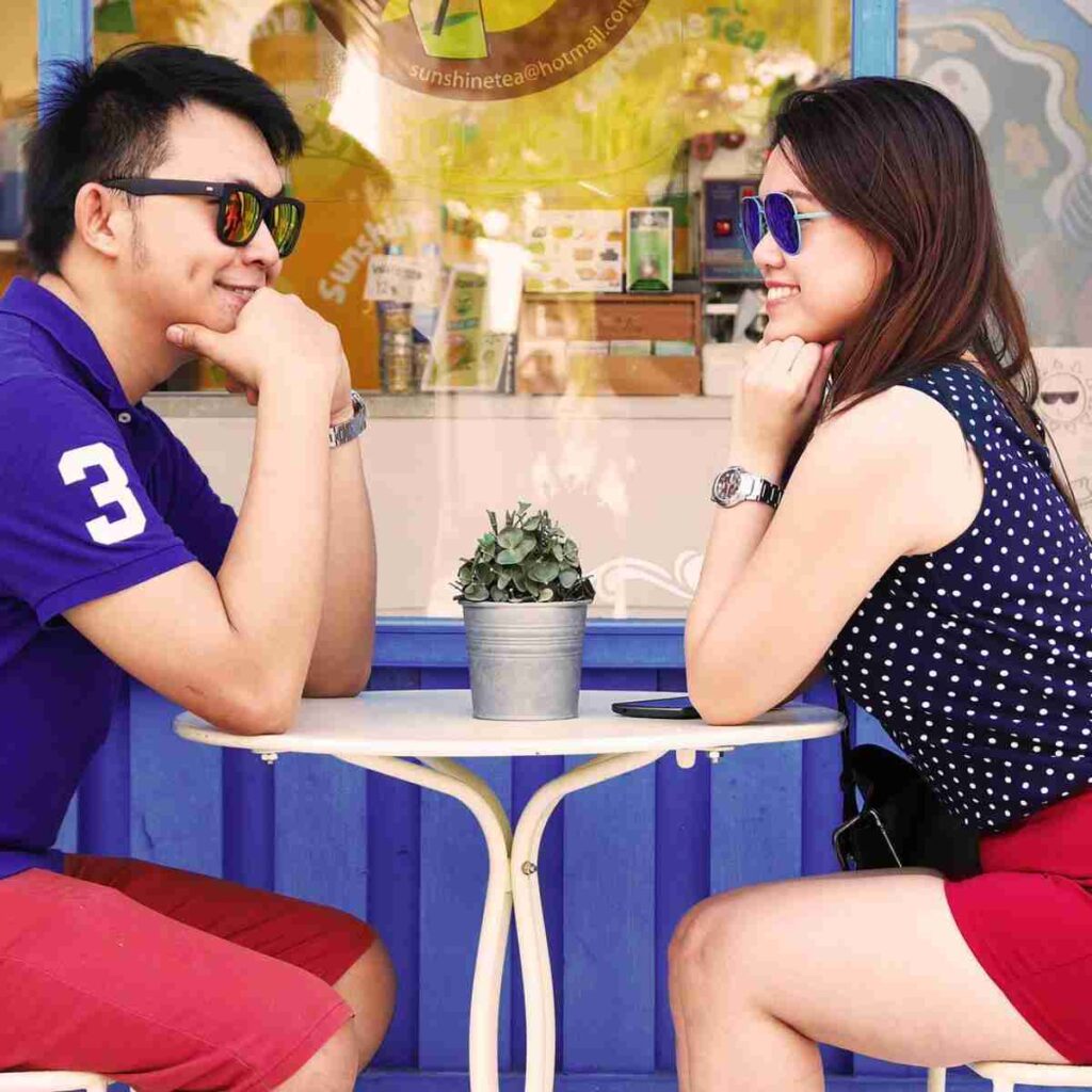 An Asian couple sits at an outdoor cafe gazing and smiling at each other.