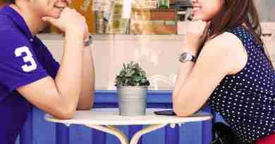 An Asian couple sits at an outdoor cafe gazing and smiling at each other.