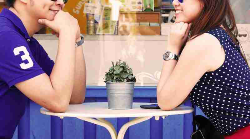An Asian couple sits at an outdoor cafe gazing and smiling at each other.