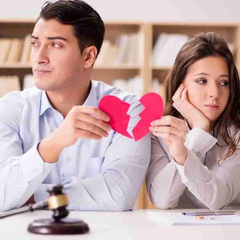 A man and a woman each look away from each other while holding a heart torn in half, signed papers, and their rings on top of them signifying their divorce.