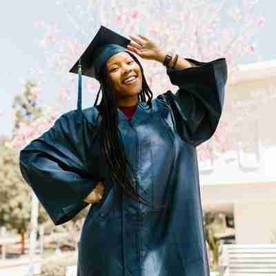 A young female graduate of color poses proudly in front of a ree in her cap and gown.