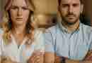 A young couple sits closely on a couch with arms crossed and serious expressions, facing forward. Both wear casual clothing—white tops and jeans—and appear to be in a moment of tension or disagreement, suggesting a relationship conflict or emotional distance.