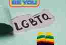 A green background with the words "Be You" and "LGBTQ" on it. There are a pair of feet wearing rainbow socks in the corner.