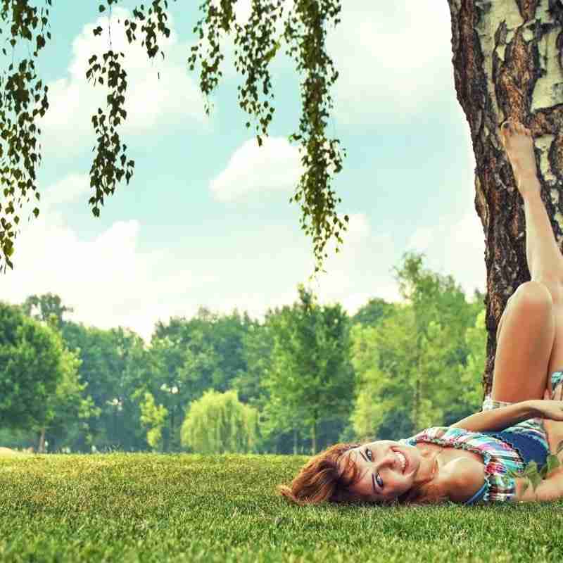 A woman laying on the grass on a bright sunny June day with her feet propped up on a tree.