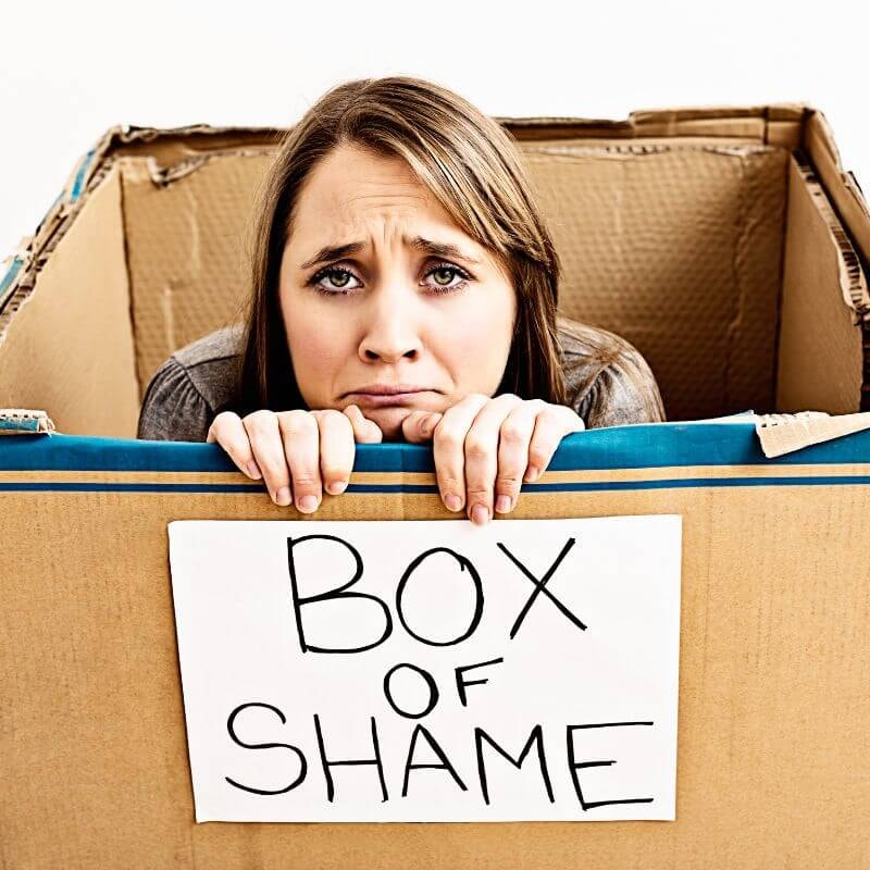 A woman sits and peers out from a card board box with a sign on it that says box of shame.