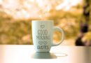 A white coffee cup with the words "Good Morning Quotes" and hearts on a wood table with trees in the background.