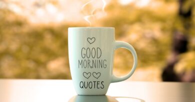A white coffee cup with the words "Good Morning Quotes" and hearts on a wood table with trees in the background.