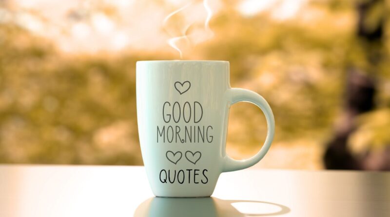 A white coffee cup with the words "Good Morning Quotes" and hearts on a wood table with trees in the background.
