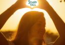 A young woman holds her hands over her head in a heart shape. The words stand tall are written inside the heart hands with the sun illuminating behind them.