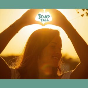 A young woman holds her hands over her head in a heart shape. The words stand tall are written inside the heart hands with the sun illuminating behind them.