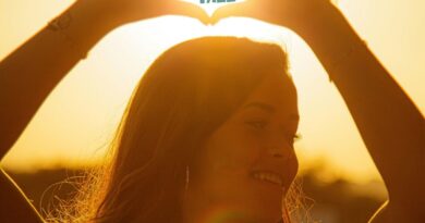 A young woman holds her hands over her head in a heart shape. The words stand tall are written inside the heart hands with the sun illuminating behind them.